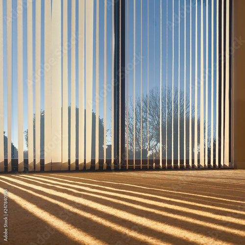sunlight through vertical blinds angled slightly open hanging over large sliding interior glass doors and throwing a striped shadow on to a beige carpet