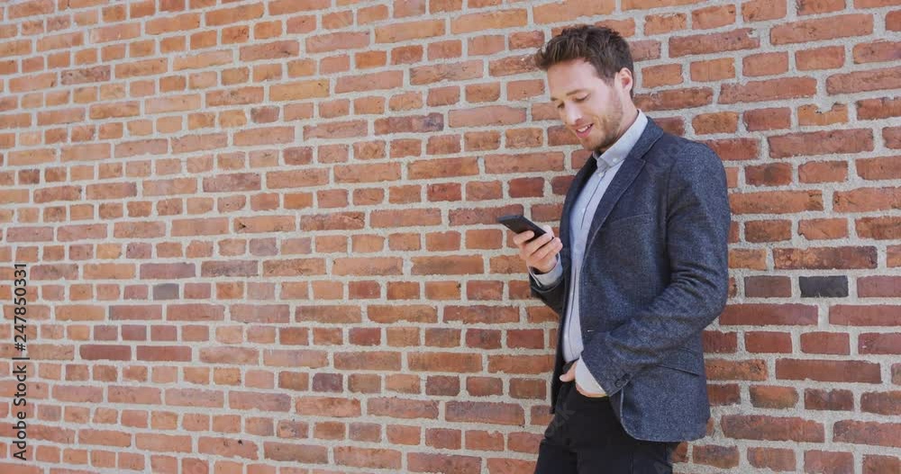 Man using phone - Smiling businessman using smartphone. Male professional standing against brick wall wearing smart casual business attire. Slow motion shot on RED. Man using phone - Smiling businessman using smartphone. Male professional standing against brick wall wearing smart casual business attire. Slow motion shot on RED.