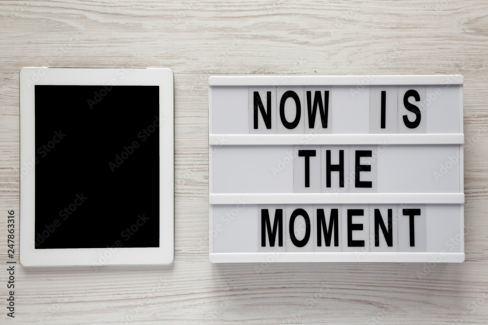 Modern board with text 'Now is the moment', tablet with blank screen on a white wooden surface, overhead view. From above, flat lay, top view.