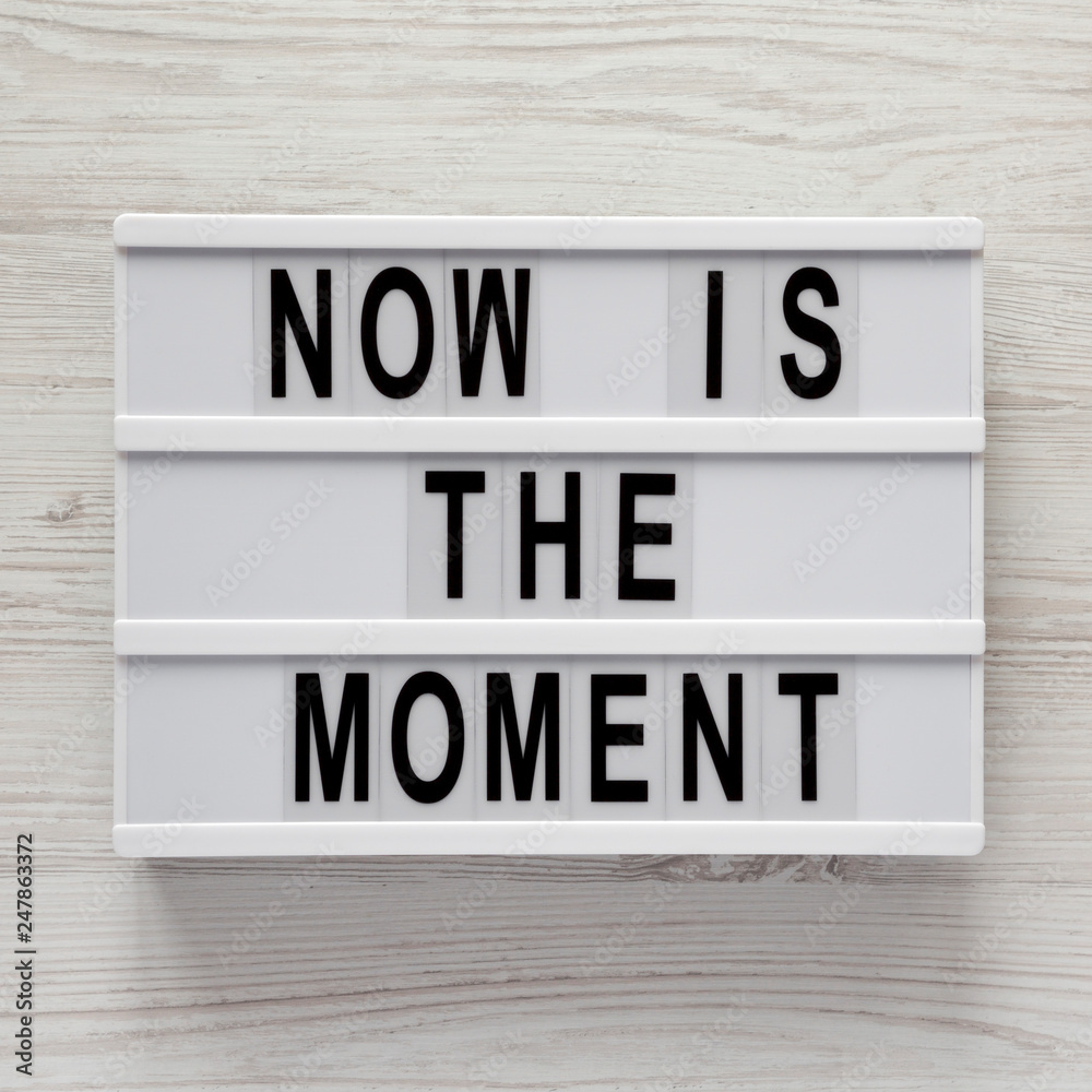 Top view, modern board with text 'Now is the moment' on a white wooden surface. From above, flat lay, overhead. Top view, modern board with text 'Now is the moment' on a white wooden surface. From above, flat lay, overhead.