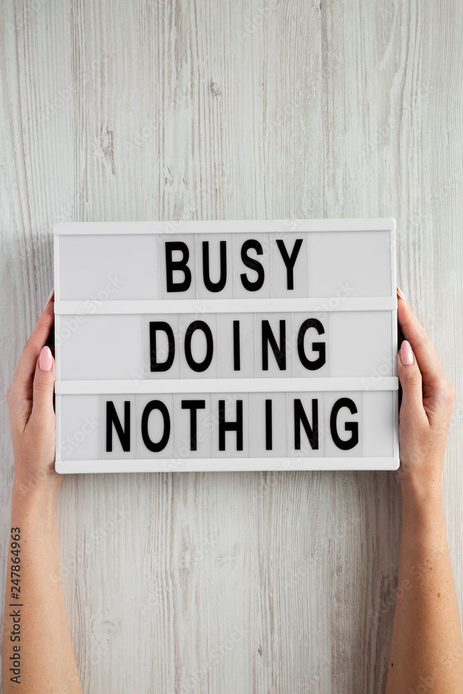 Female hands hold lightbox with text 'Busy doing nothing' on a white wooden table, top view. From above, flat lay, overhead. Female hands hold lightbox with text 'Busy doing nothing' on a white wooden table, top view. From above, flat lay, overhead.