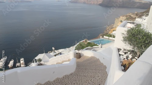 Looking down at the cliffs and holiday villas that engulf the Aegean sea in Oia, Santorini. Tranquille sea during sunrise with stairs climbing down a Greek village alley in the foreground. photo