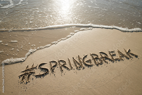 Modern Spring Break message handwritten with a social media hashtag on the smooth sand of an empty beach with an oncoming wave