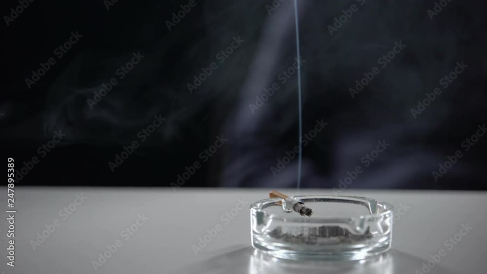 Cigarette burning in ash tray isolated on dark background, nicotine addiction Cigarette burning in ash tray isolated on dark background, nicotine addiction