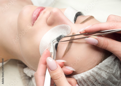 Eyelash extension procedure. Woman face and experts fingers with tweezers, close up, selective focus