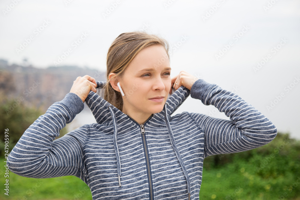 Serious young woman putting hoodie hood on outdoors