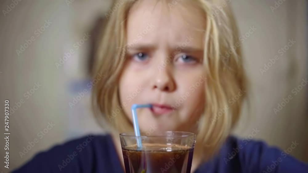 pretty preschooler girl is blowing through plastic straw into glass with soda lemonade, making bubbles