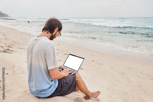 A young man works at the laptop on the beach. Freelancer working on the beach by the ocean.