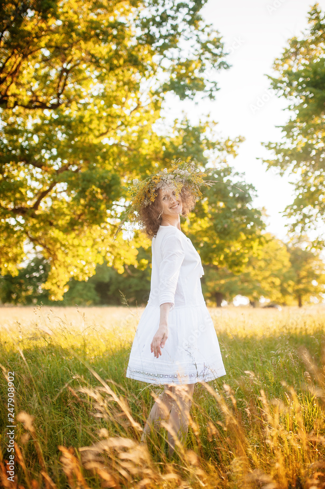 Caucasian happy girl in a white dress with a wreath of wild flowers in the summer at sunset in the field.