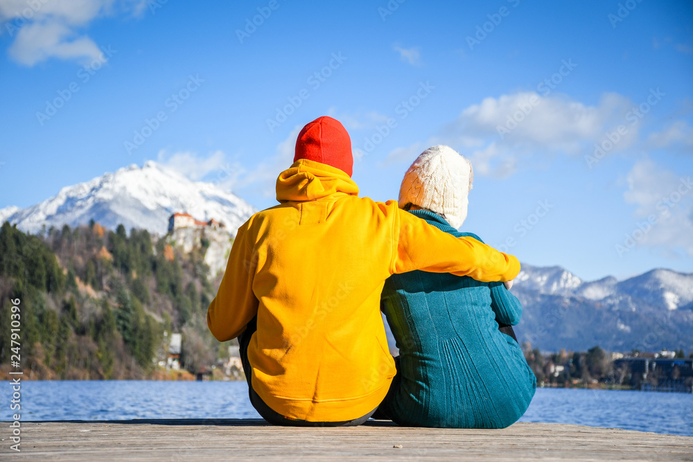 Fototapeta premium Couple in love hugging together with colorful cloths sitting and relaxing on a wooden pier on a clear sky sunny winter day view from the back, Bled