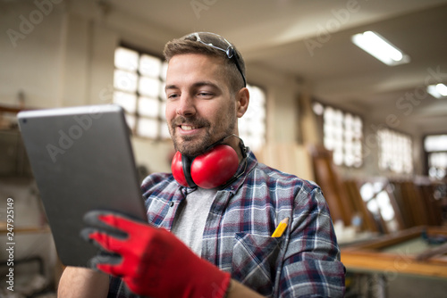 Cuadro en lienzo Portrait of middle aged professional worker carpenter with ear protectors using tablet in carpentry workshop