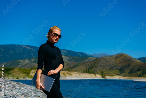Freelancer with laptop on the nature. Blogger hipster traveler business lady pretty girl finished work outdoor. Young blondy woman with sunglasses in black standing next to mountain river