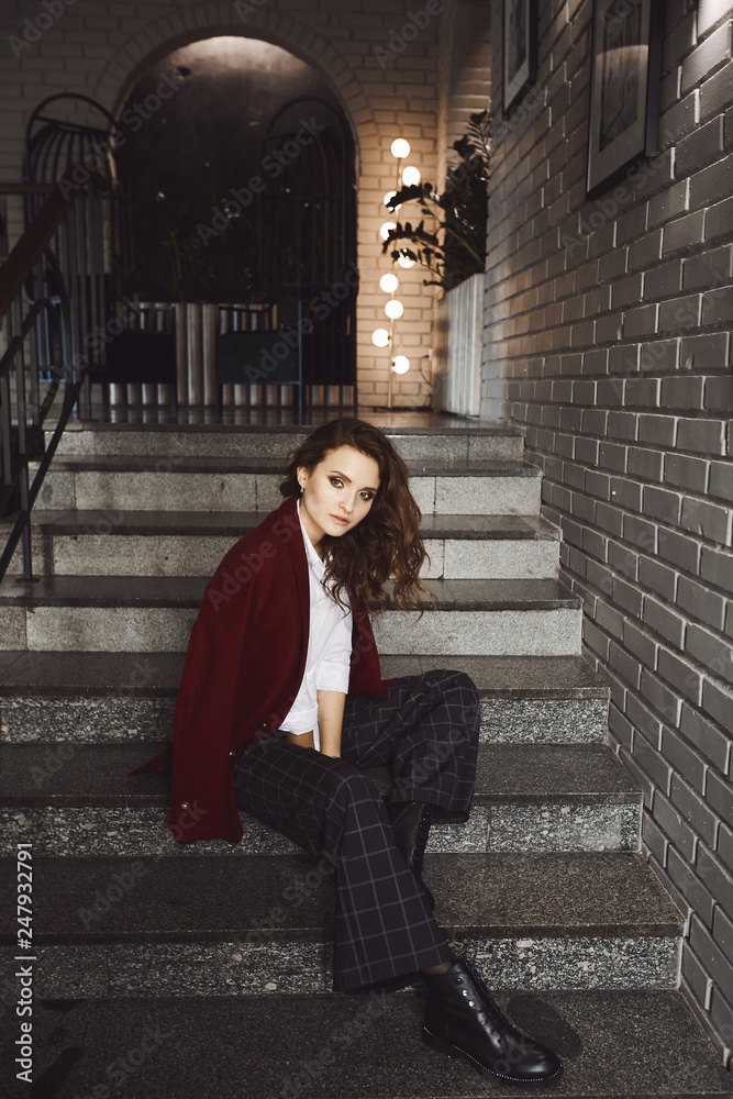 Beautiful and fashionable brunette model girl with perfect makeup in a stylish red jacket and in trendy plaid pants sits on the stairs and posing at loft interior