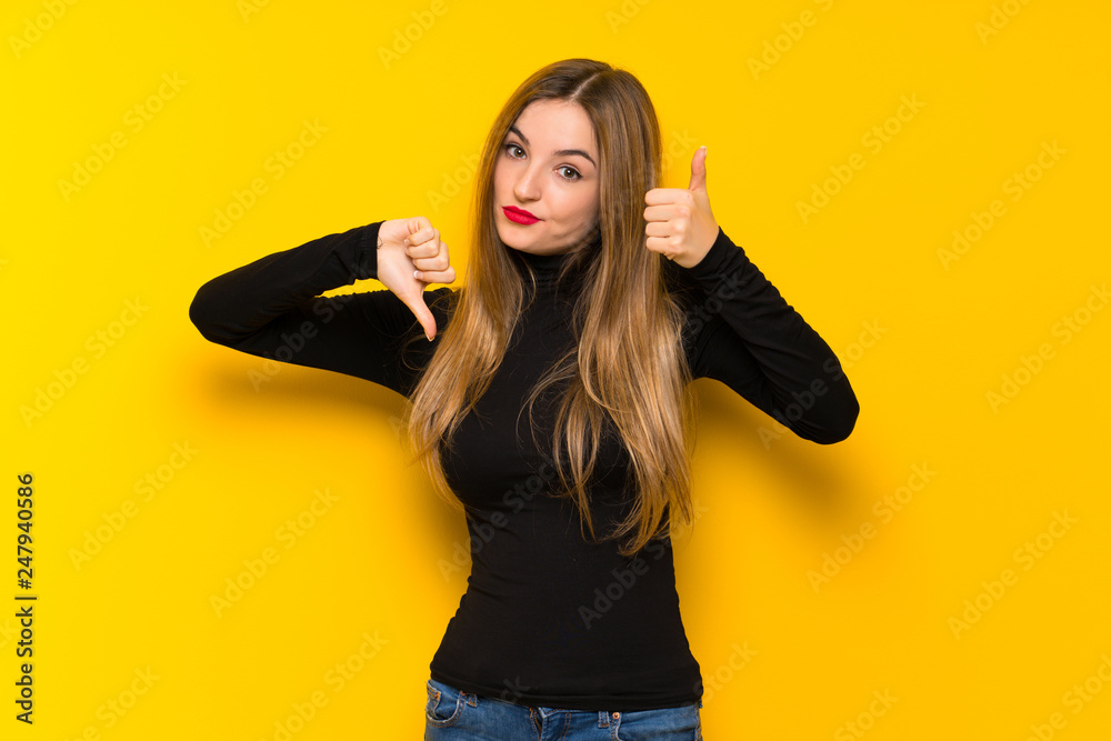 Young pretty woman over yellow background making good-bad sign. Undecided between yes or not Young pretty woman over yellow background making good-bad sign. Undecided between yes or not