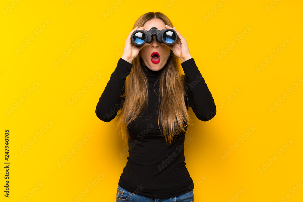 Young pretty woman over yellow background and looking in the distance with binoculars - obrazy, fototapety, plakaty 