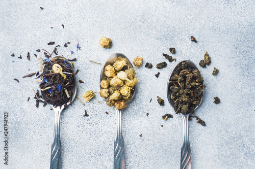 Variety of dry tea leaves and flowers in spoons on grey background
