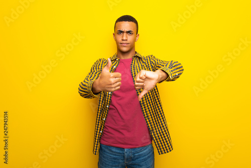 Young afro american man on yellow background making good-bad sign. Undecided between yes or not