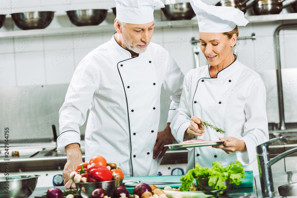 female and male chefs in double-breasted jackets and hats decorating dish with rosemary while cooking in restaurant kitchen female and male chefs in double-breasted jackets and hats decorating dish with rosemary while cooking in restaurant kitchen