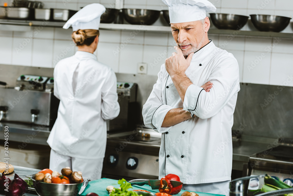 pensive male chef in double-breasted jacket during cooking in restaurant kitchen with female colleague on background