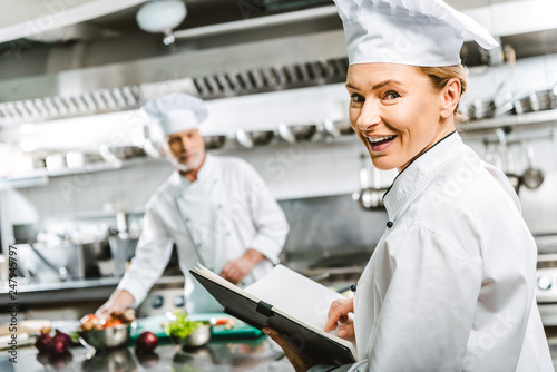 beautiful female chef in uniform holding recipe book and looking at camera in restaurant kitchen with colleague on background