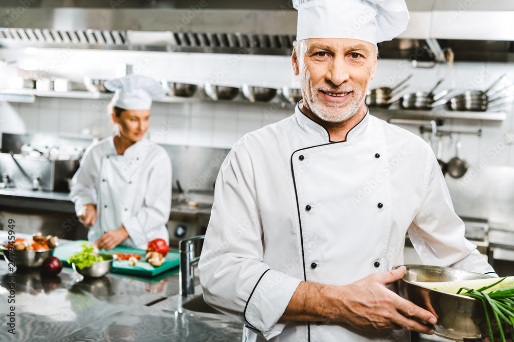 handsome male chef in uniform looking at camera while colleague cooking on background in restaurant kitchen