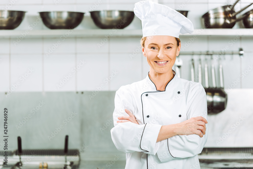 beautiful smiling female chef in uniform and hat with arms crossed looking at camera in restaurant kitchen