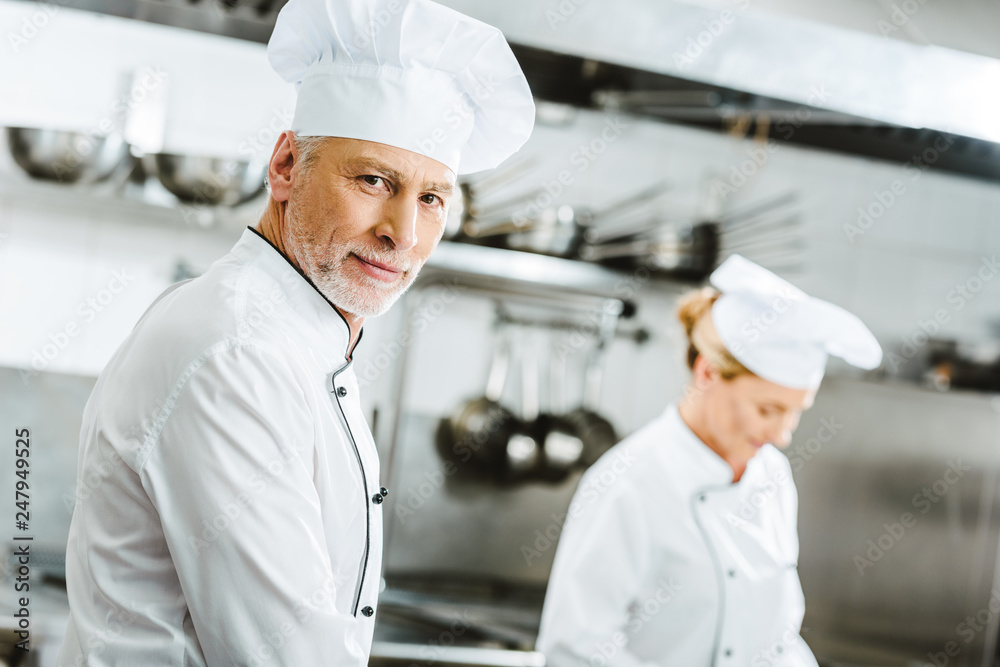 selective focus of handsome male chef in double-breasted jacket and hat looking at camera in restaurant kitchen