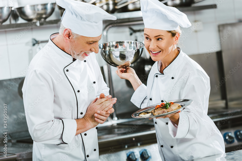 female chef in uniform presenting meat dish on serving tray to male cook in restaurant kitchen