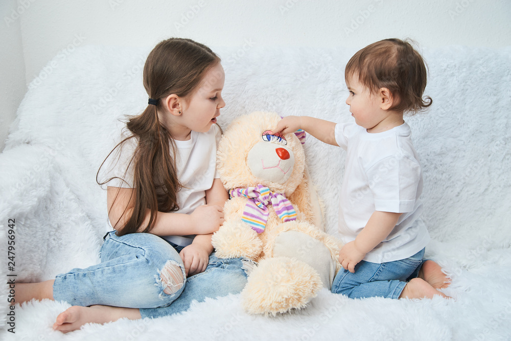 Two children, sisters play on a white sofa in white t-shirts and blue jeans. Soft plush Bunny Two children, sisters play on a white sofa in white t-shirts and blue jeans. Soft plush Bunny