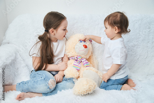 Two children, sisters play on a white sofa in white t-shirts and blue jeans. Soft plush Bunny