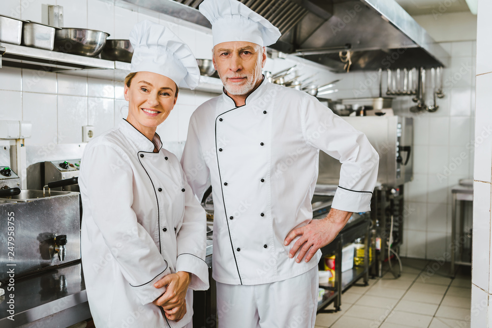 female and male chefs in uniform looking at camera and smiling at restaurant kitchen
