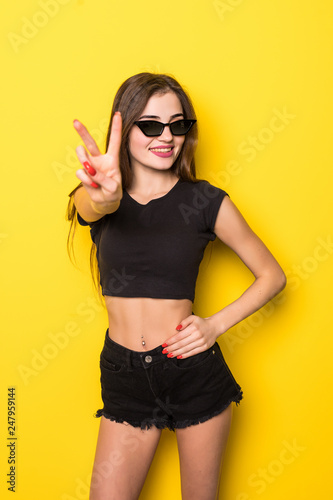 Portrait of happy young woman showing peace gesture standing isolated over yellow background