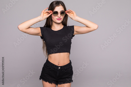 Portrait of young woman holding hands on sunglasses wearing in summer outfit isolated on gray background