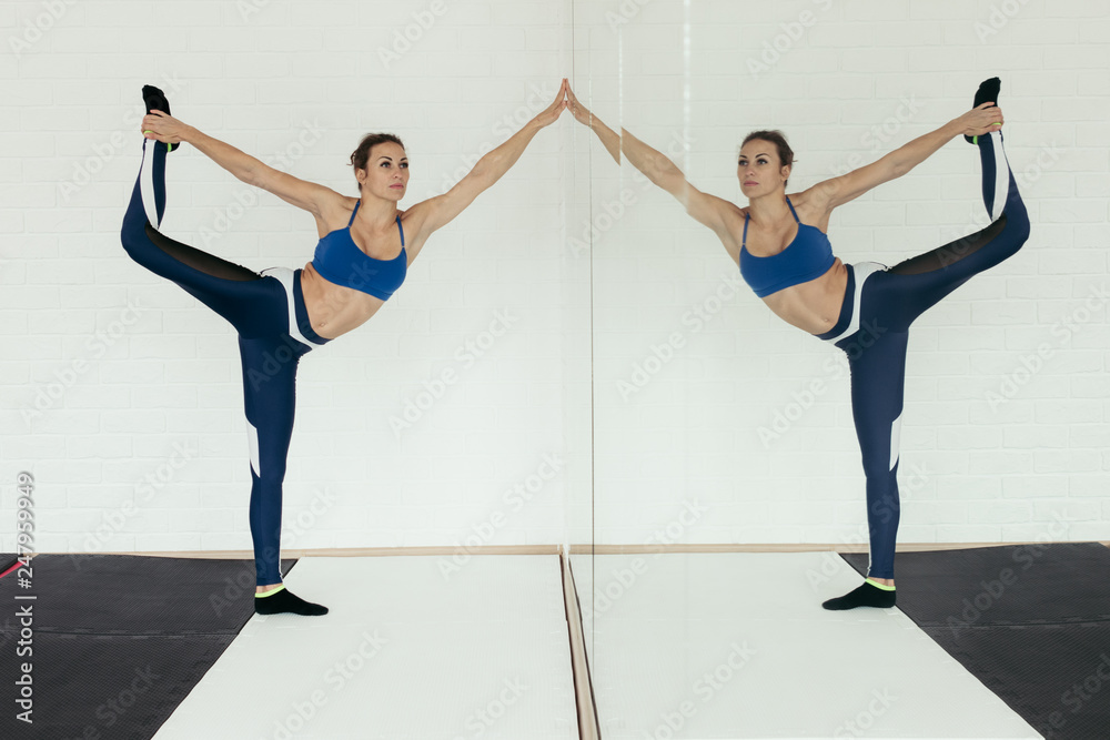 Naklejka premium Dark hair girl makes yoga in front of mirror placed in white wall studio