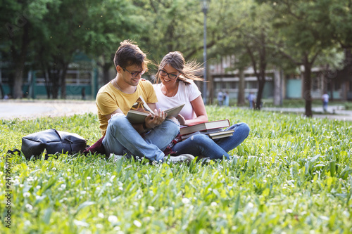 Two young students sitting at the campus yard , reading book and preparing for university exam.