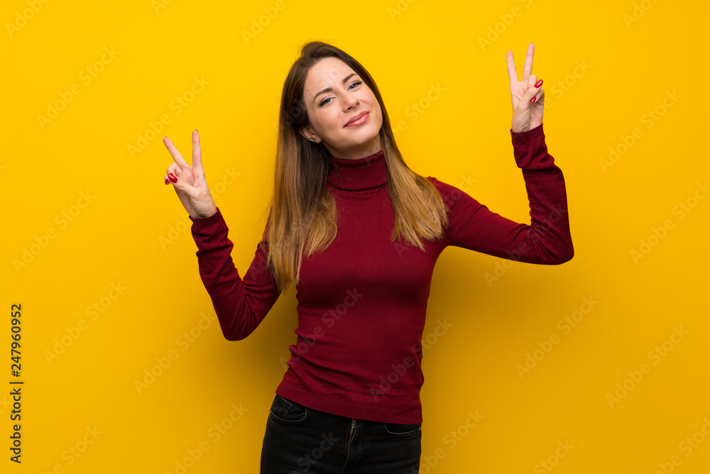 Woman with turtleneck over yellow wall smiling and showing victory sign with both hands Woman with turtleneck over yellow wall smiling and showing victory sign with both hands