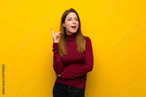 Woman with turtleneck over yellow wall thinking an idea pointing the finger up