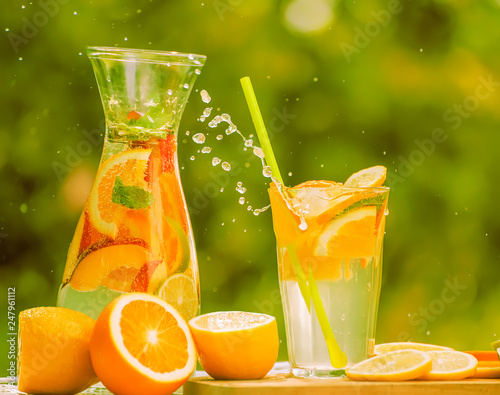 A carafe and a glass with an illuminating summer drink  lemonade and various fruits against the backdrop of a green garden.