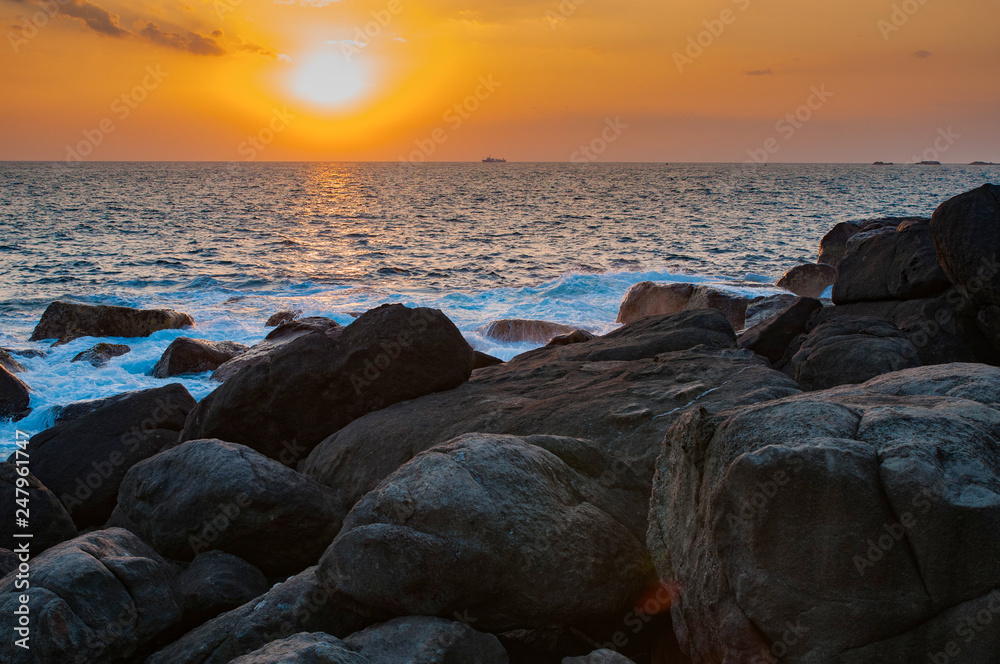 Sunset over the sea with ship on horizon and waves near the rocks.