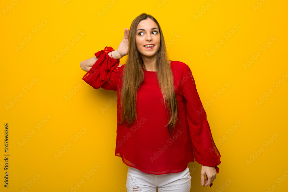 Young girl with red dress over yellow wall listening to something by putting hand on the ear Young girl with red dress over yellow wall listening to something by putting hand on the ear