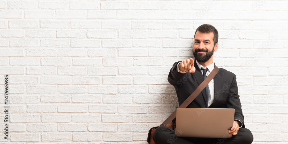 Businessman with his laptop sitting on the floor points finger at you with a confident expression Businessman with his laptop sitting on the floor points finger at you with a confident expression