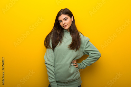 Teenager girl with green sweatshirt on yellow background posing with arms at hip and smiling