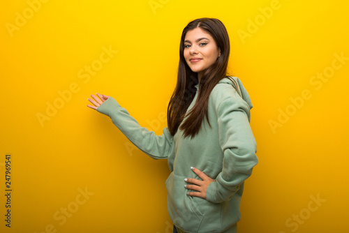Teenager girl with green sweatshirt on yellow background pointing back and presenting a product