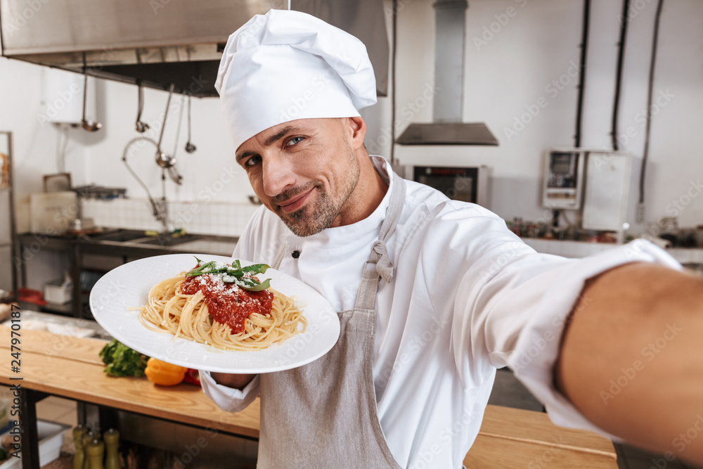 Image of handsome man chief in apron taking selfie, while standing at kitchen in restaurant Image of handsome man chief in apron taking selfie, while standing at kitchen in restaurant