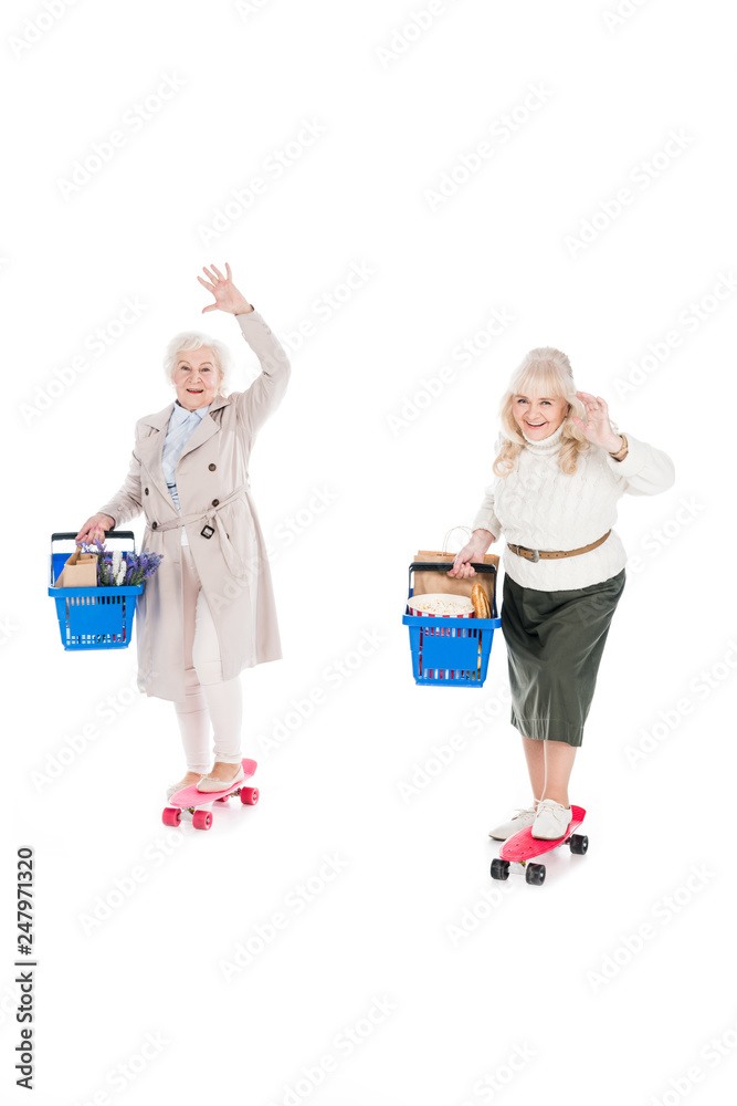 happy senior women riding penny boards and holding shopping baskets isolated on white