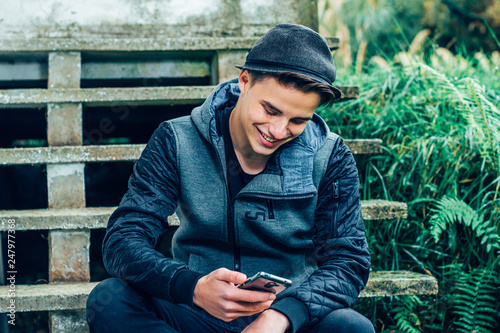 Happy young man wearing a stylish hat sitting outdoors and smiling while looking at mobile phone.