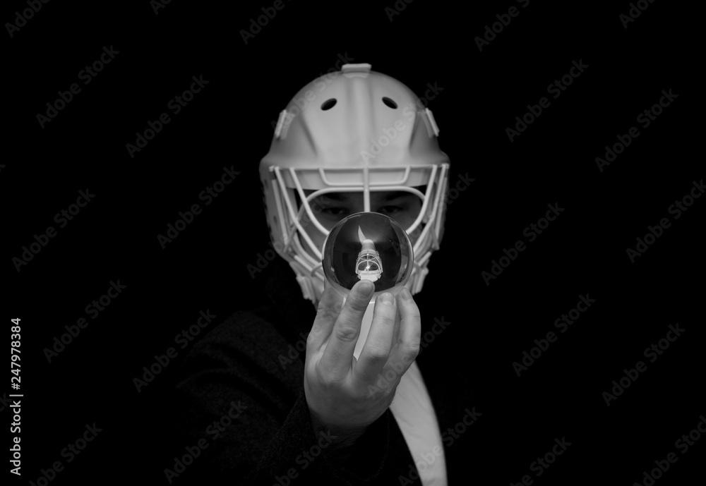 A young man has a goalie helmet on his head and holds a crystal ball with his reflection.Black and white photo.Man in a jacket with a goalie helmet on his head estimates the outcome of the match.