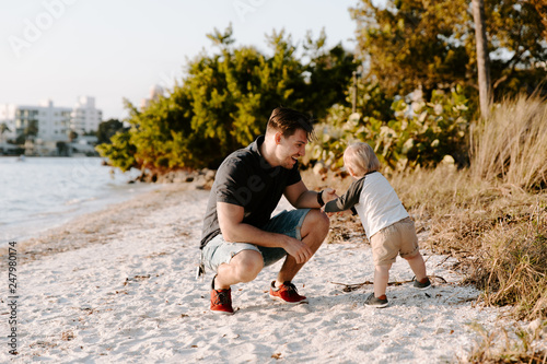 Young Father and His Little Boy Son Walking and Enjoying the Nice Outdoor Weather on the Sandy Beach next to the Ocean Bay with Sail Boats in the Background