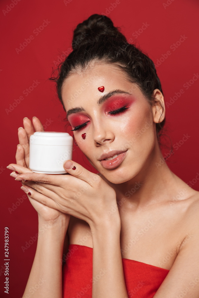 Beautiful young woman with red bright makeup isolated over red wall background posing with hearts on face holding cream. Beautiful young woman with red bright makeup isolated over red wall background posing with hearts on face holding cream.