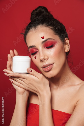 Beautiful young woman with red bright makeup isolated over red wall background posing with hearts on face holding cream.